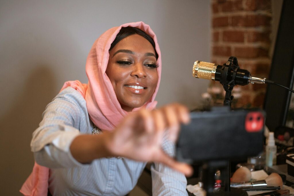 A Black woman in a hijab selfies while vlogging with a microphone indoors.